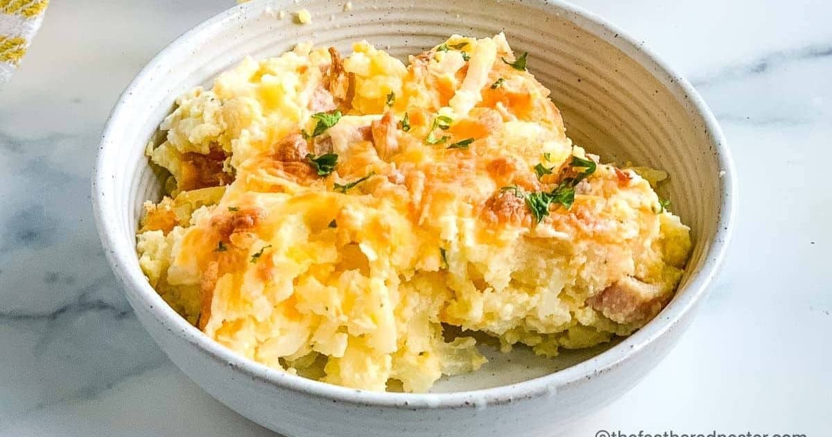 A bowl of cheesy baked mashed potatoes garnished with chopped herbs on a marble surface.