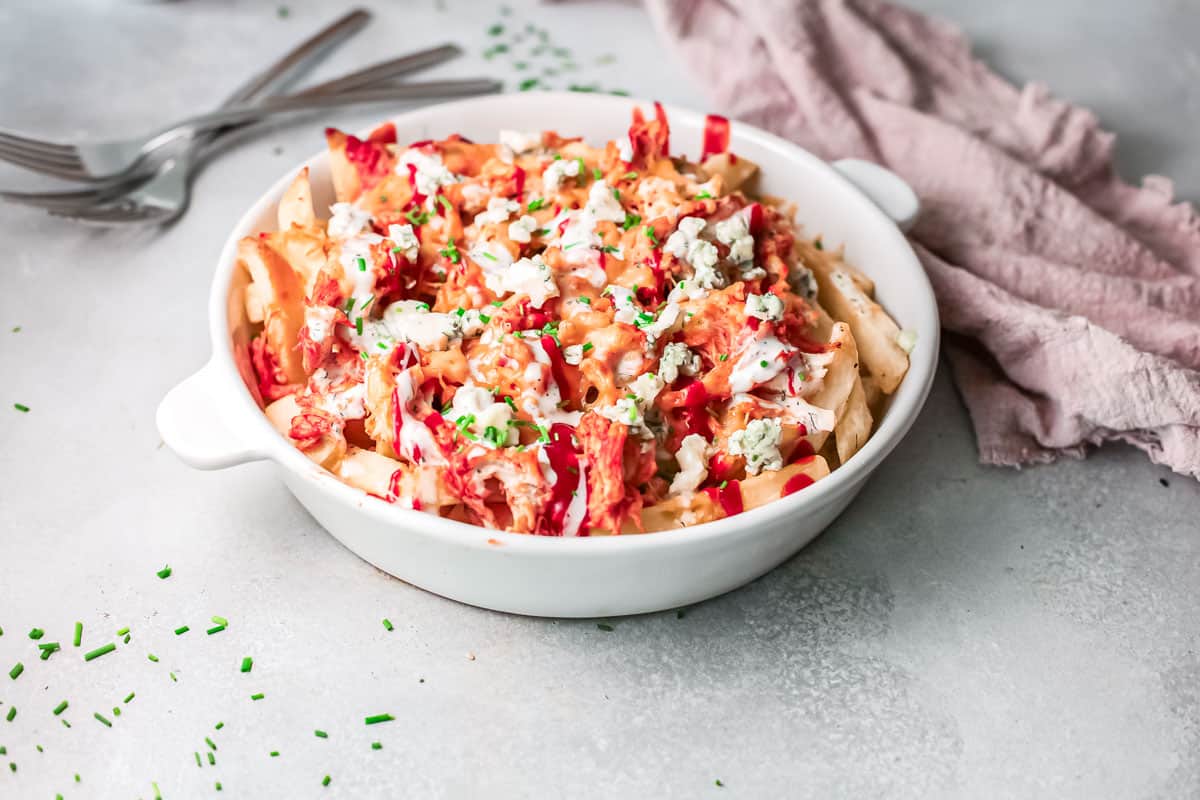 White bowl of loaded fries topped with cheese, chives, and sauce, next to a pink cloth and forks.