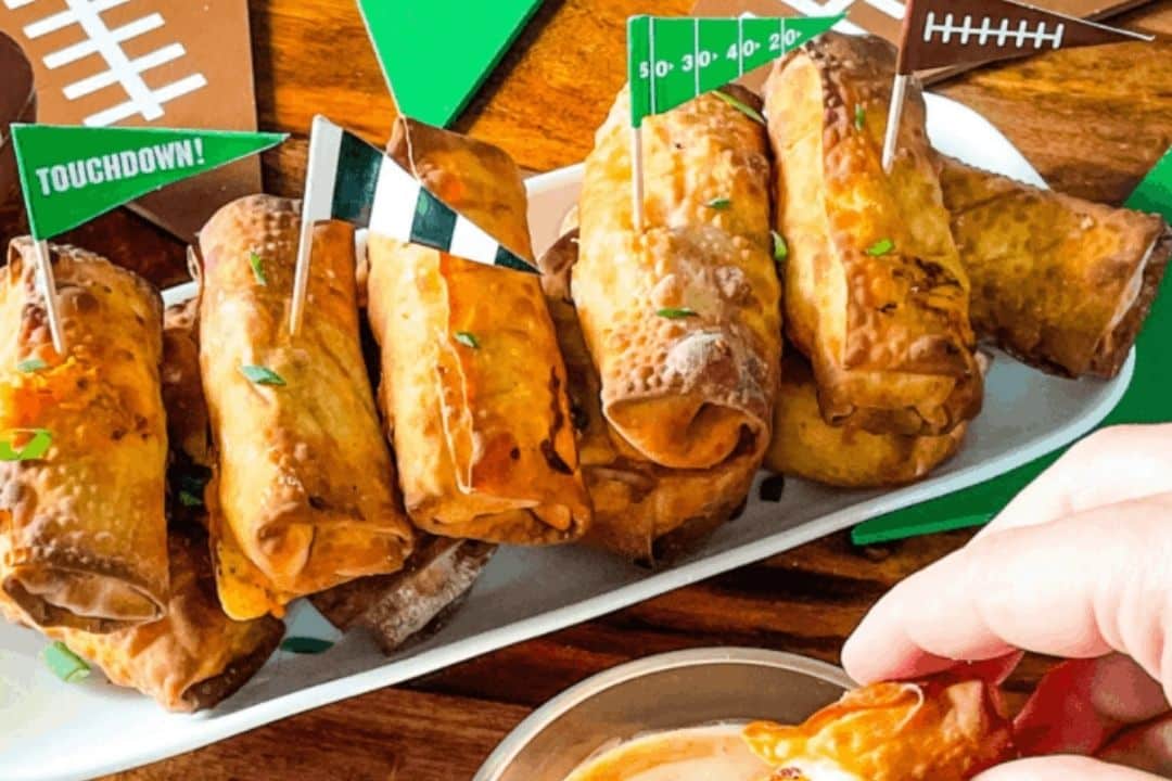 Plate of crispy egg rolls with football-themed flags; a hand dips one into sauce in the foreground.