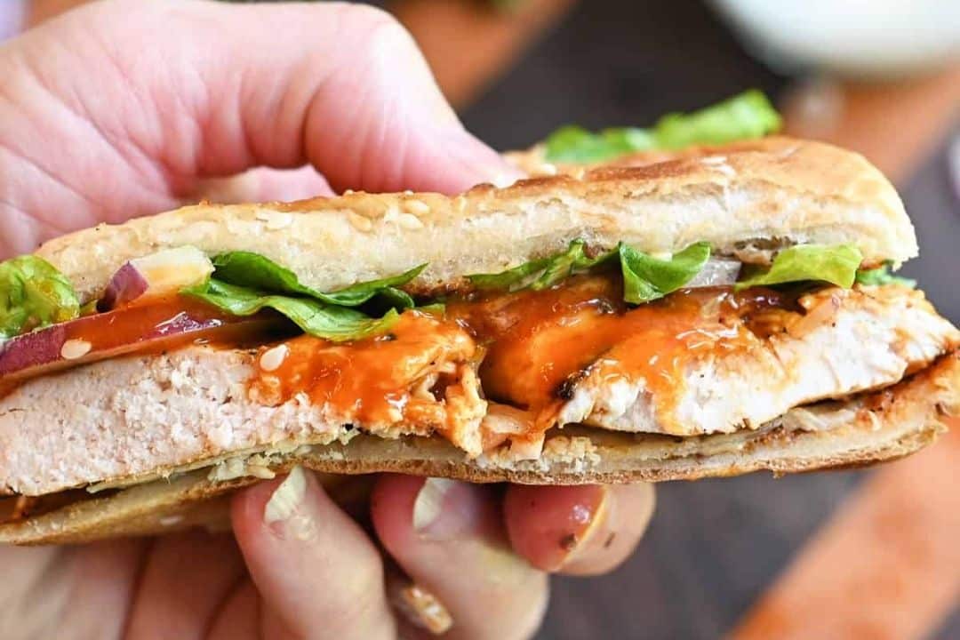 A hand holding a sesame sandwich with grilled chicken, lettuce, onions, and a tangy orange sauce.