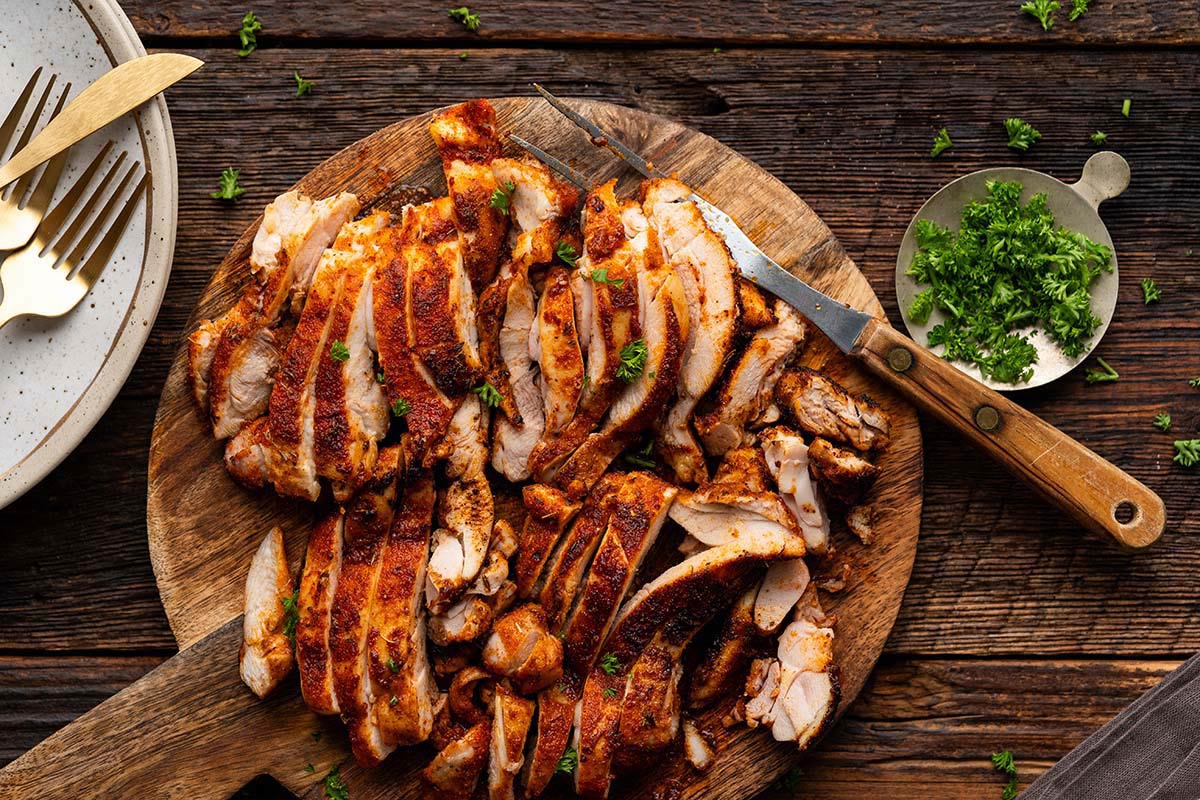 Sliced grilled chicken with herbs on a wooden board, knife beside, parsley garnish on the side.