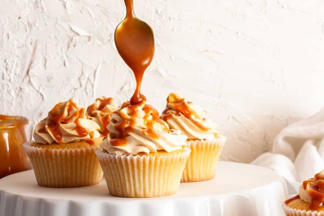 Caramel being drizzled over cupcakes with swirled frosting, displayed on a white cake stand.