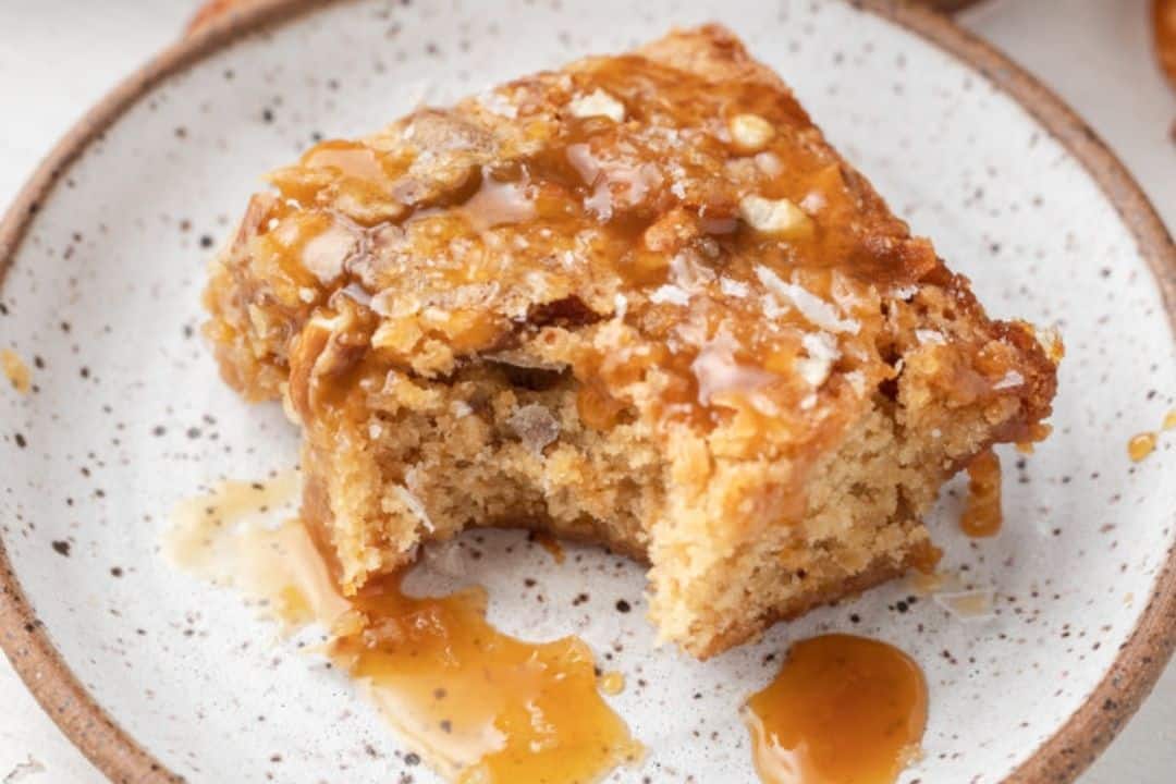 A square piece of cake with caramel sauce on a plate, with a bite taken out of it.