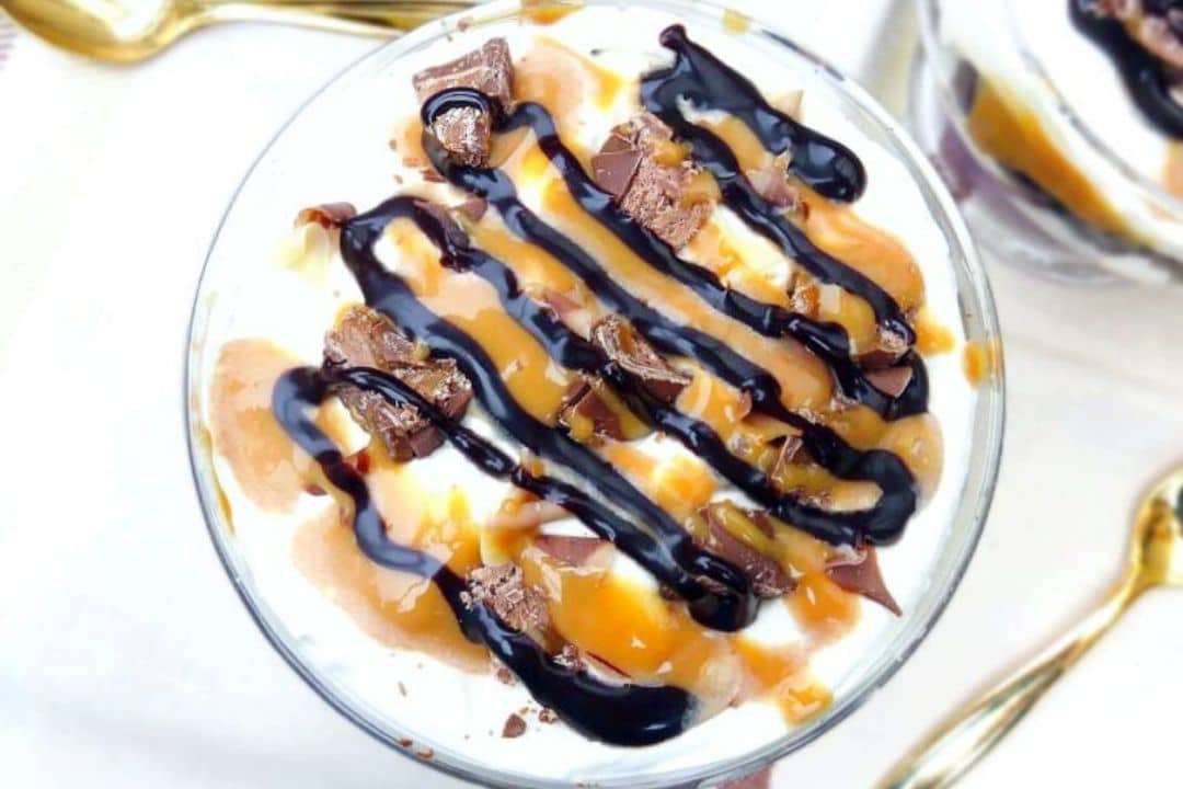 A dessert topped with caramel, chocolate sauce, and chocolate pieces in a glass dish with a spoon nearby.