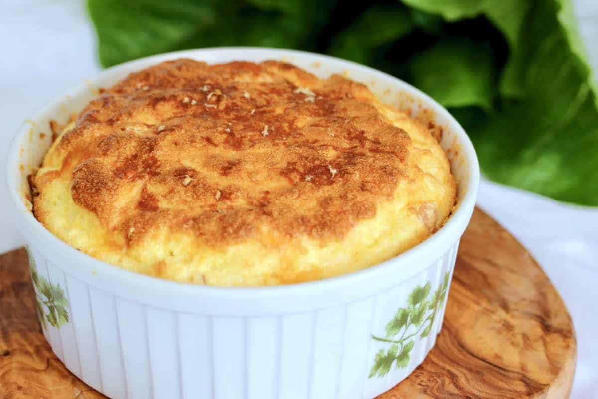 Golden-brown cheese soufflé in a white ramekin with green leaf pattern, placed on a wooden board.