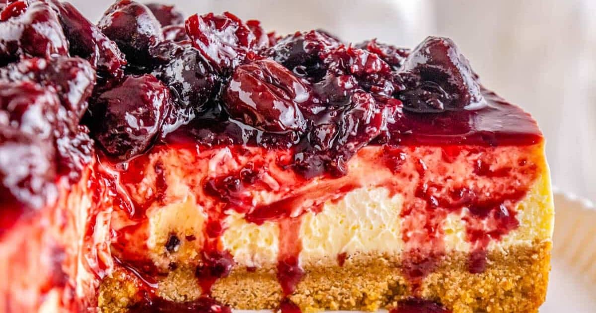 A slice of cheesecake topped with glossy cherry sauce and whole cherries, showing a crumbly cookie crust.
