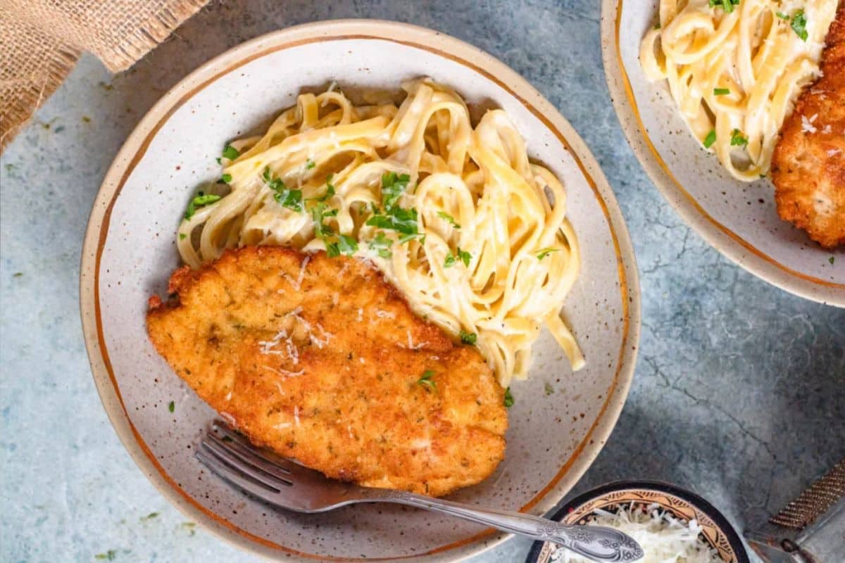 A plate of breaded chicken cutlet with creamy fettuccine pasta, garnished with chopped herbs.