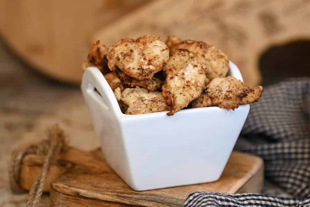 Golden brown fried chicken bites in a white square bowl on a wooden board, with a checkered cloth nearby.