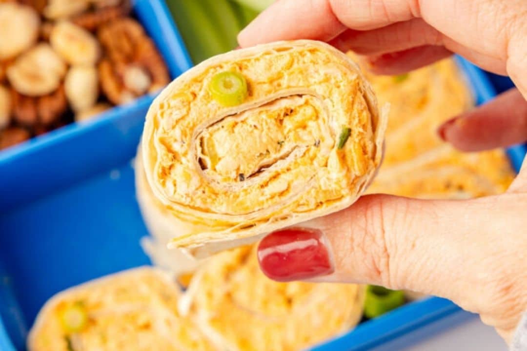 A hand holding a wrap filled with a creamy, orange mixture, with a lunchbox and nuts in the background.