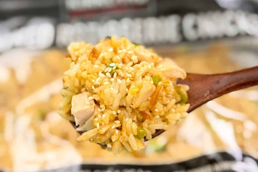 A wooden spoon holds fried rice with vegetables, chicken pieces, and sprinkled sesame seeds.