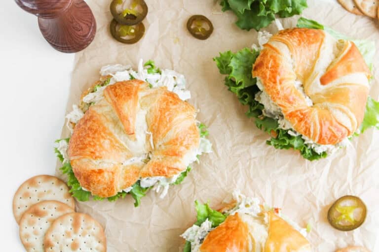 Three croissant sandwiches with lettuce and chicken salad on parchment paper, with crackers and jalapeño slices.