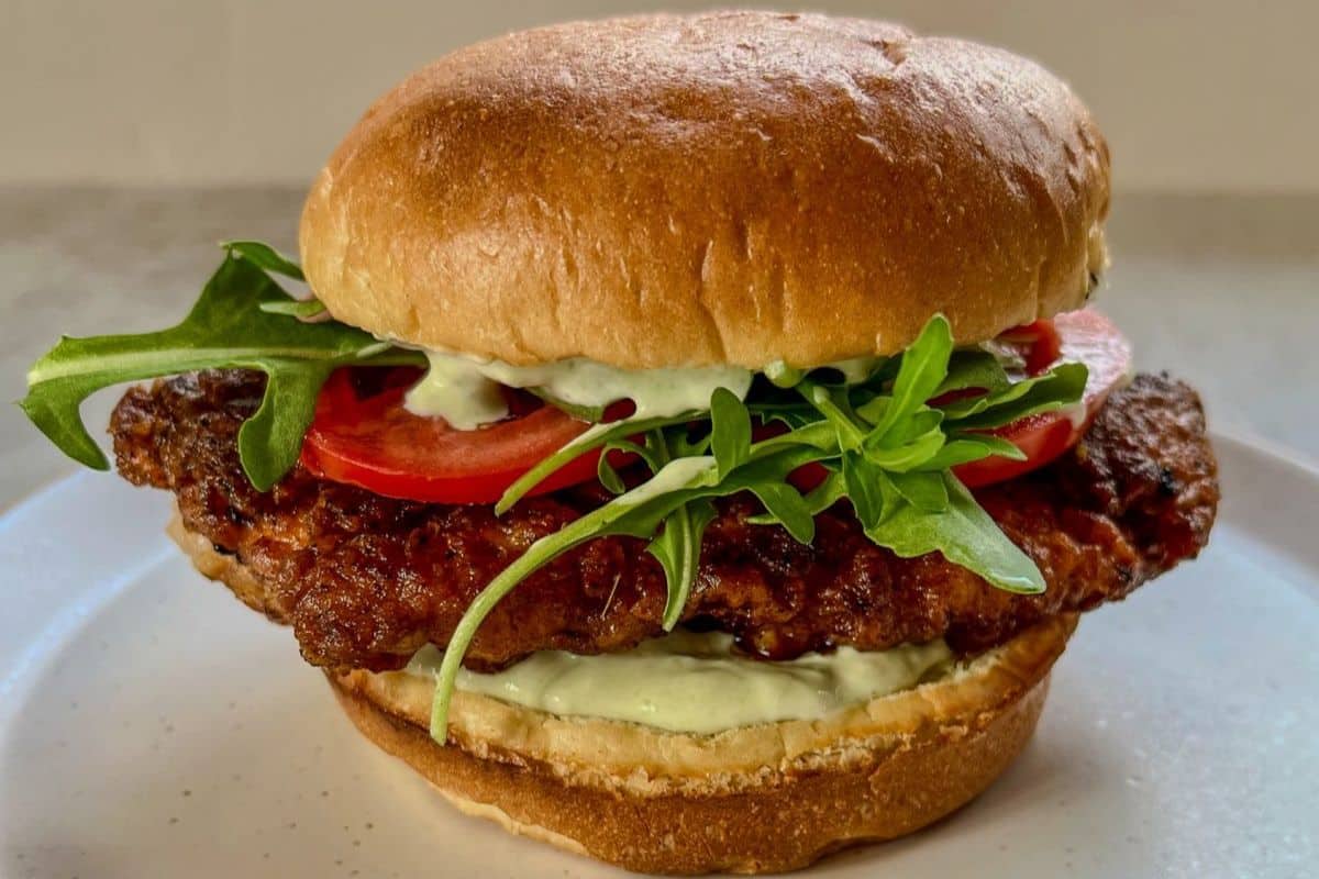 Crispy chicken sandwich with tomato, arugula, and sauce on a toasted bun, served on a white plate.