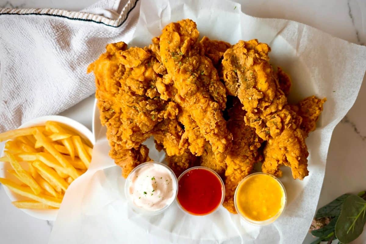 A plate of crispy fried chicken with three dipping sauces and a side of French fries on a white table.