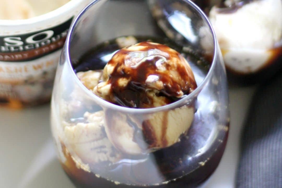 A glass with ice cream scoops topped with chocolate syrup and a tub of ice cream in the background.