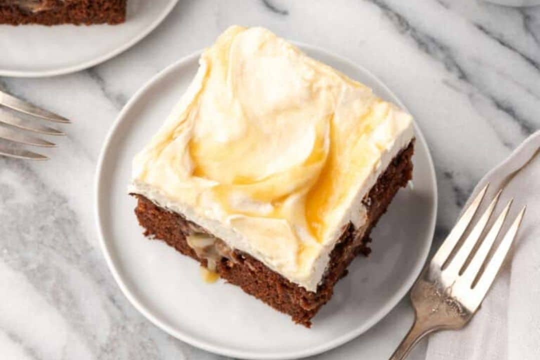 A slice of chocolate cake with creamy, swirled frosting on a plate, next to a fork on a marble surface.