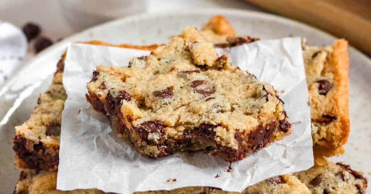 A close-up of chocolate chip cookie bars stacked on a plate, with one bar resting on parchment paper.