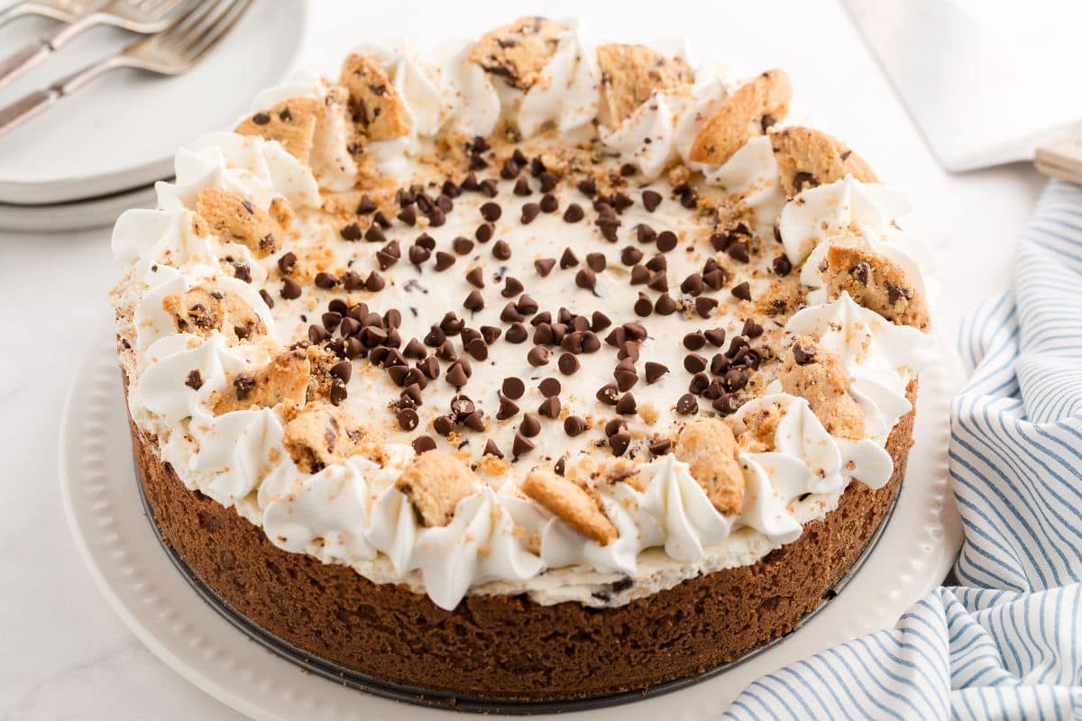 A cookie cake topped with whipped cream, chocolate chips, and cookie pieces on a white plate.