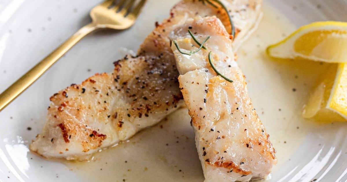 Two pieces of cooked white fish with pepper, sauce, rosemary, lemon wedges, and a gold fork on a white plate.