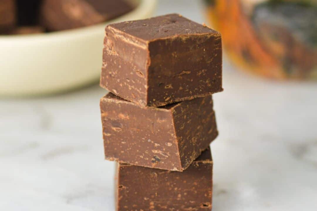 Three pieces of chocolate fudge stacked on a white surface, with a bowl in the background.