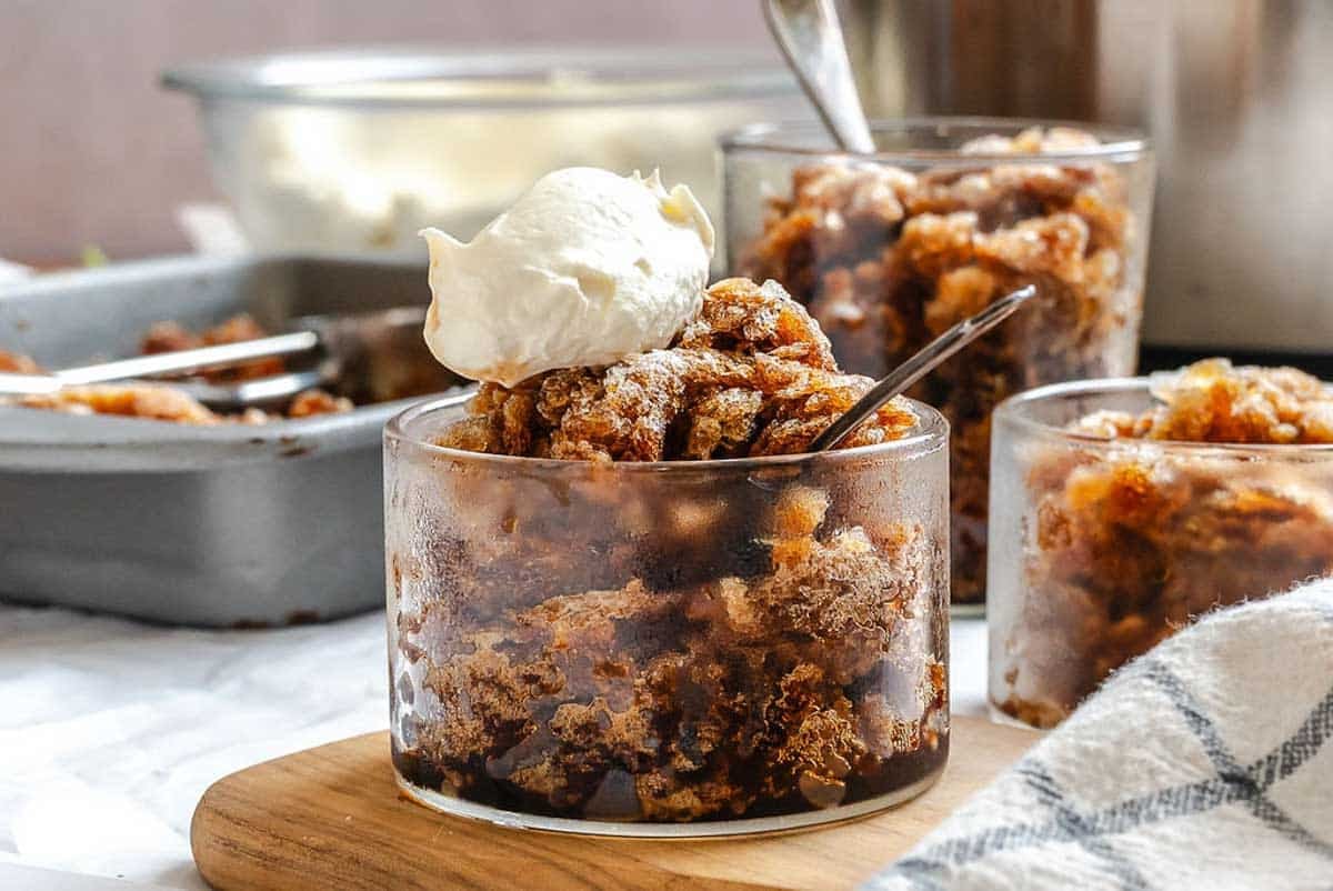 A glass of spiced bread pudding topped with whipped cream, with more servings in the background.
