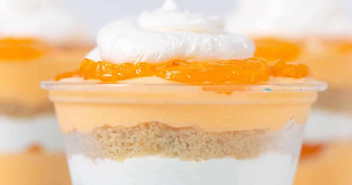 A layered dessert cup with whipped cream, orange topping, crumb layer, and white creamy base.