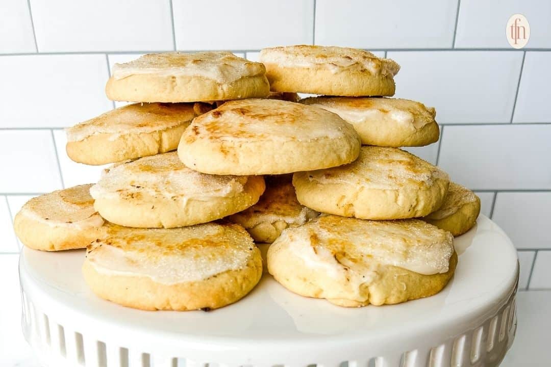 A stack of frosted sugar cookies on a white cake stand in front of a tiled wall.