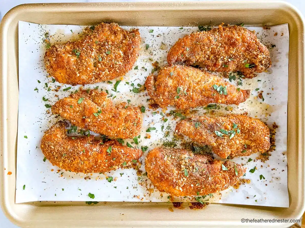 Eight breaded chicken tenders baked on a parchment-lined baking sheet, garnished with chopped herbs.