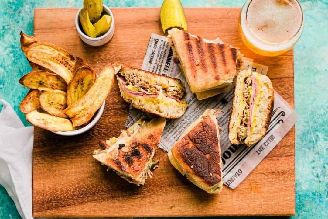 Grilled sandwiches, fried plantains, pickles, and a drink on a wooden board with newspaper-style paper.