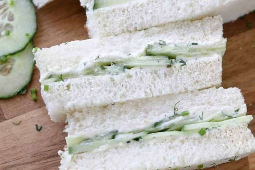 Three cucumber tea sandwiches with white bread and cream filling on a wooden surface, next to cucumber slices.