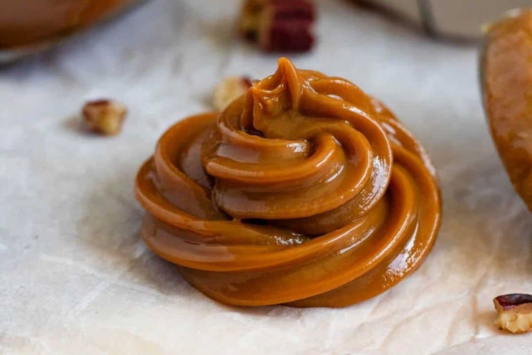 A swirl of creamy caramel sauce sits on parchment paper with a few nut pieces nearby.