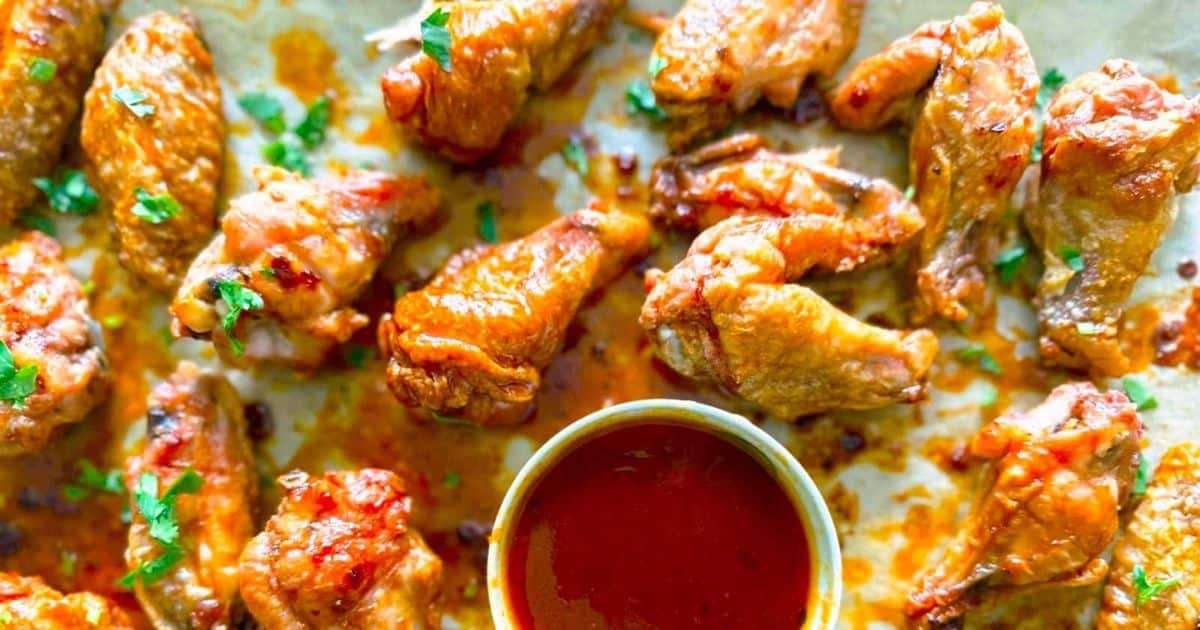 Crispy baked chicken wings with chopped herbs, served with a bowl of red dipping sauce on a tray.