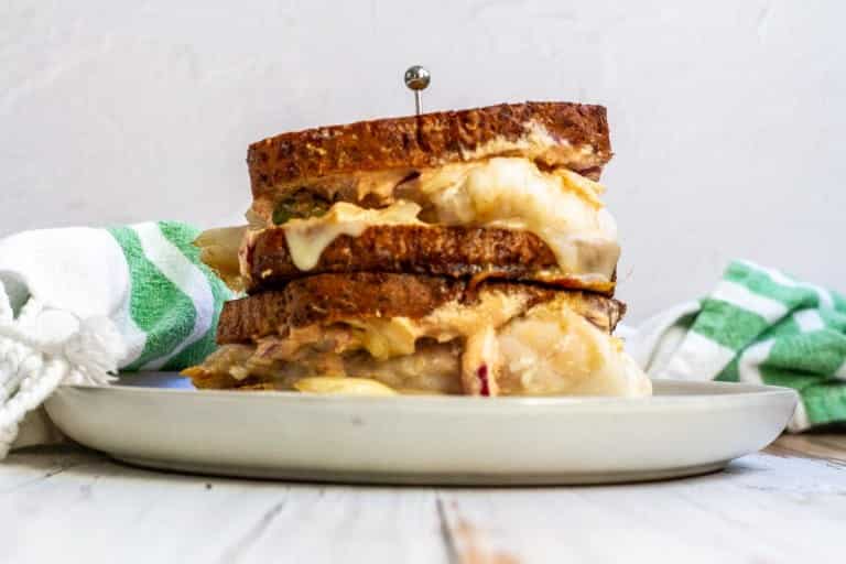 A stacked grilled sandwich with melted cheese on a plate, secured with a toothpick, next to a green-striped towel.