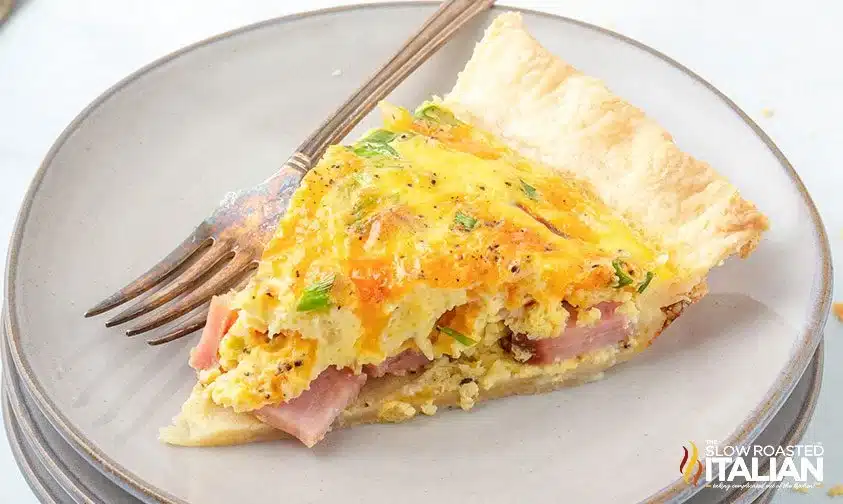 A slice of ham and cheese quiche on a plate with a fork.
