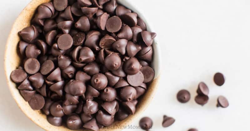 A bowl filled with dark chocolate chips, with a few chips scattered on a white surface nearby.