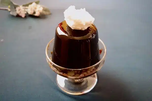 A glass dessert cup with coffee jelly topped with whipped cream, set on a blue surface.