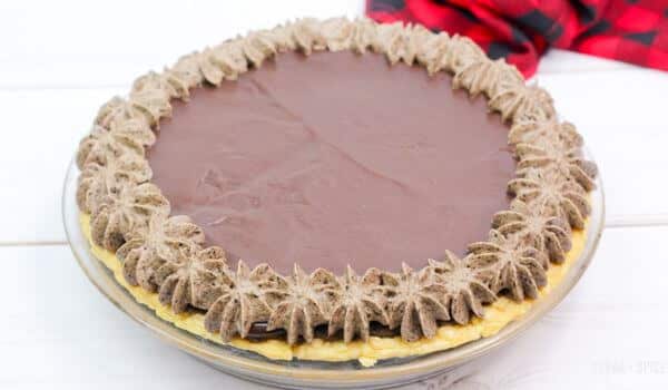 Chocolate cream pie with whipped topping piped around the edge, on a light surface with a red plaid cloth nearby.