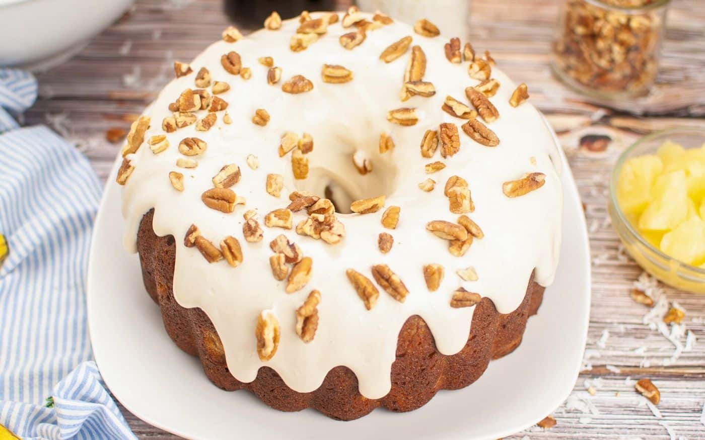 Bundt cake with white icing, topped with chopped pecans, on a white plate on a wooden table.