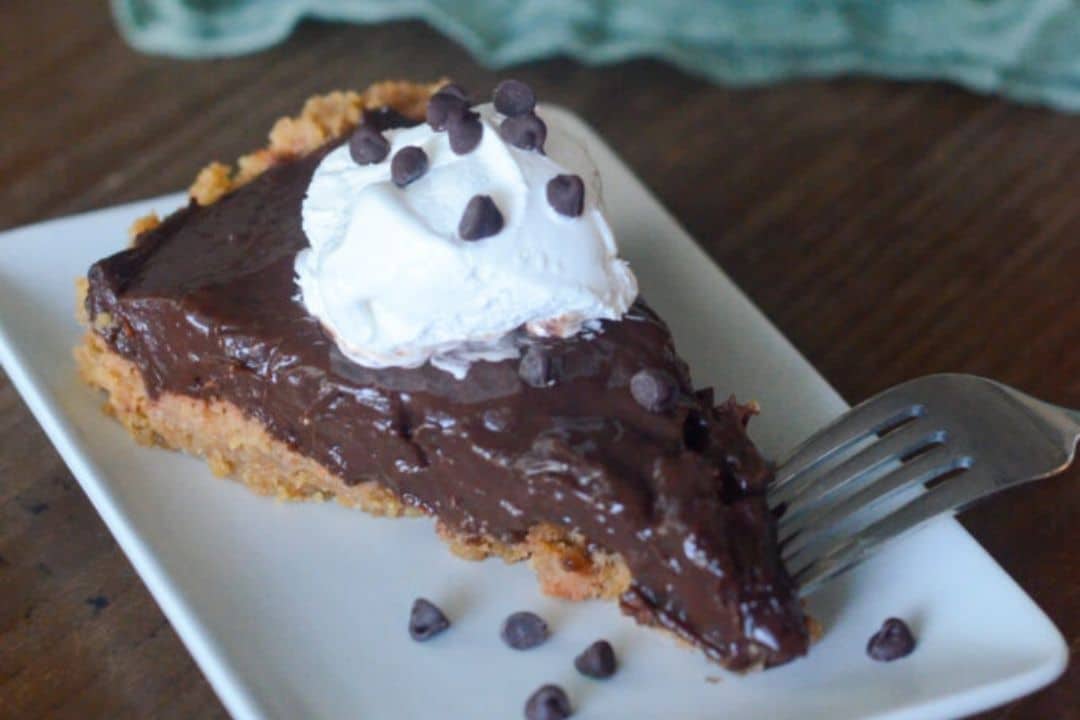 A slice of chocolate pie topped with whipped cream and chocolate chips on a white plate with a fork.
