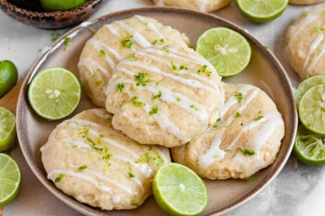 Four iced lime cookies on a plate, garnished with lime zest and surrounded by fresh lime halves.