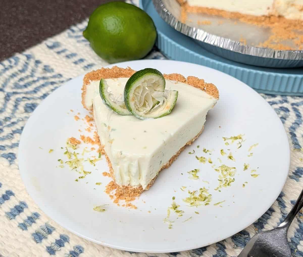 A slice of key lime pie garnished with lime, on a white plate with lime zest, next to a whole lime.