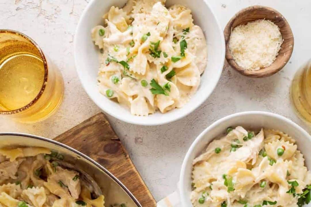 Bowls of creamy pasta with peas, a glass of drink, grated cheese in a small bowl, and a pan on the table.