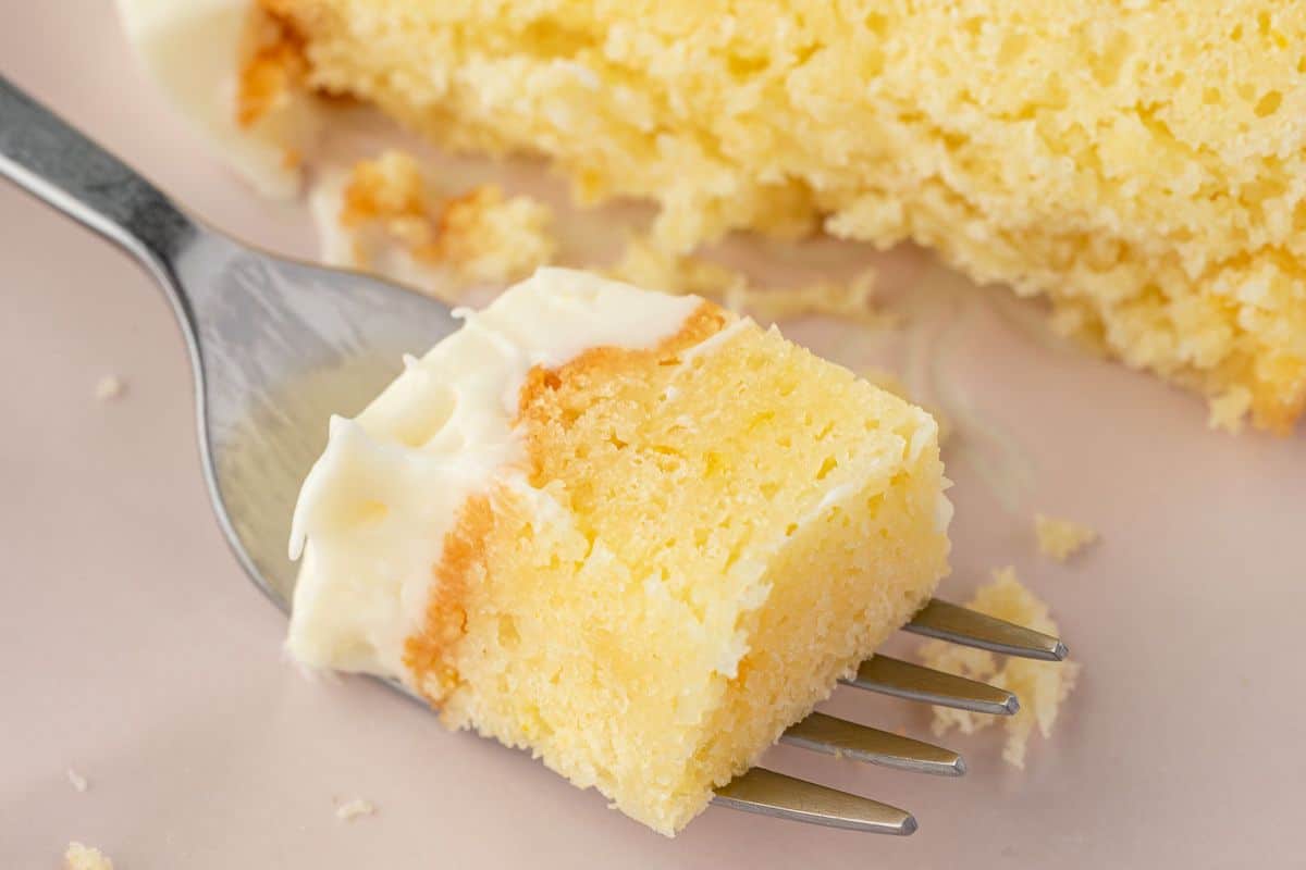 A fork holding a bite of moist yellow cake with white frosting, with more cake in the background.