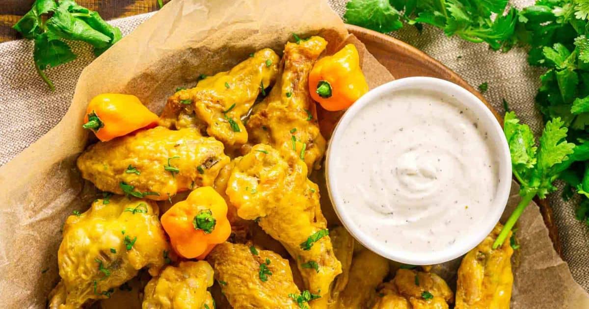 Chicken wings coated in yellow sauce, garnished with herbs and peppers, served with a bowl of creamy dipping sauce.
