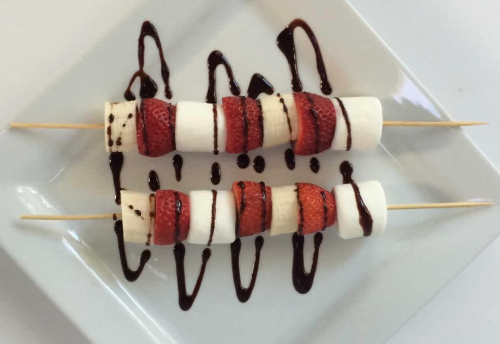 Two skewers with marshmallows, strawberries, and bananas, drizzled with chocolate sauce, on a white plate.