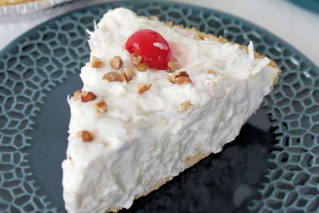 A slice of creamy pie topped with chopped nuts, shredded coconut, and a red cherry on a dark blue plate.