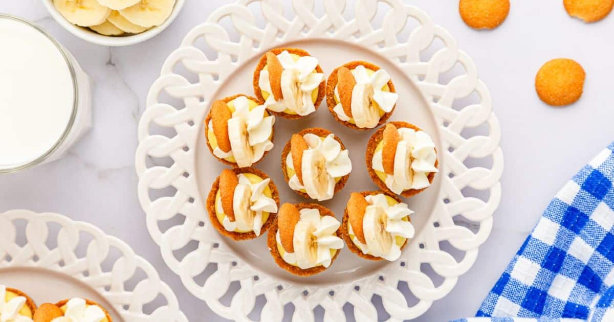 Round cookies with cream and banana slices on a white plate, next to a blue checkered cloth and a glass of milk.