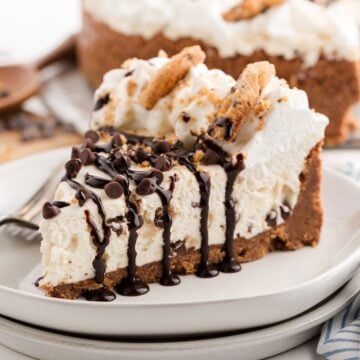 A slice of creamy cheesecake with chocolate chip cookie crust, whipped cream, and chocolate drizzle on a plate.