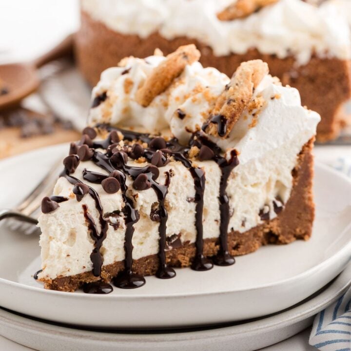 A slice of creamy cheesecake with chocolate chip cookie crust, whipped cream, and chocolate drizzle on a plate.