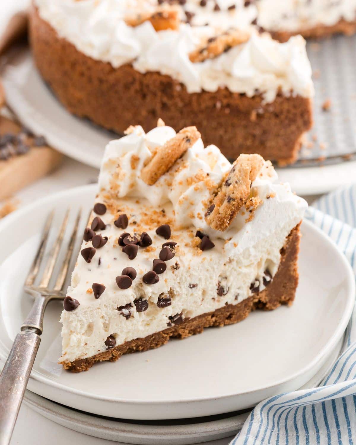 Slice of creamy pie topped with whipped cream, chocolate chips, and cookie pieces on a white plate with a fork.