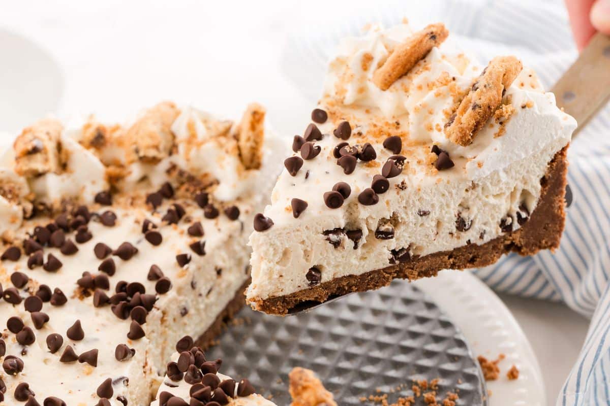 A slice of creamy pie topped with whipped cream, mini chocolate chips, and cookie pieces being served.
